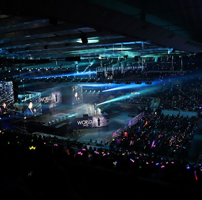 A large indoor arena filled with an audience watching a live performance. The stage features large screens displaying a performer and graphics, with bright spotlights and ambient lighting creating a dynamic atmosphere. The crowd is holding glow sticks, adding vibrant colors to the scene.
