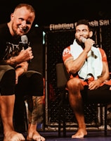 Two individuals are seated on a stage, each holding a microphone and smiling. They are in a sports-related setting with one wearing a casual black outfit and the other in sportswear, with a towel around his neck. The background includes elements of a fighting or martial arts cage.