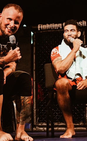 Two individuals are seated on a stage, each holding a microphone and smiling. They are in a sports-related setting with one wearing a casual black outfit and the other in sportswear, with a towel around his neck. The background includes elements of a fighting or martial arts cage.