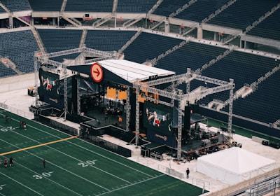 A large outdoor stage is set up inside a stadium, equipped with tall scaffolding, lights, and multiple large display screens. The seats in the stadium are mostly empty. The field is marked with numbers and lines typical of a sports venue. A few people are visible near the stage and on the field.