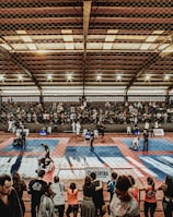 A large indoor arena with a high ceiling, filled with an audience seated on bleachers. In the center, two fighters are engaged in a martial arts competition on blue and red mats. The scene is captured through a mesh fence, adding a textured foreground.