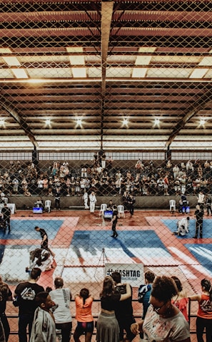 A large indoor arena with a high ceiling, filled with an audience seated on bleachers. In the center, two fighters are engaged in a martial arts competition on blue and red mats. The scene is captured through a mesh fence, adding a textured foreground.