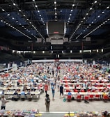 An indoor sports arena filled with rows of tables, each occupied by people engaged in competitive activities. The ceiling is high, with numerous lights illuminating the large space. The audience seating on either side is empty, and there are flags hanging from the upper level.