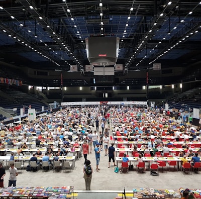 An indoor sports arena filled with rows of tables, each occupied by people engaged in competitive activities. The ceiling is high, with numerous lights illuminating the large space. The audience seating on either side is empty, and there are flags hanging from the upper level.