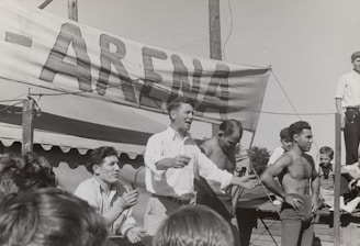 A group of men, some shirtless, are gathered around a platform or stage. A man in a white shirt appears to be speaking or gesturing to a crowd. A large sign with the word 'ARENA' is prominently displayed in the background. The scene seems to be set outdoors on a sunny day.