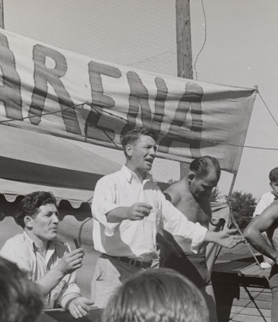 A group of men, some shirtless, are gathered around a platform or stage. A man in a white shirt appears to be speaking or gesturing to a crowd. A large sign with the word 'ARENA' is prominently displayed in the background. The scene seems to be set outdoors on a sunny day.