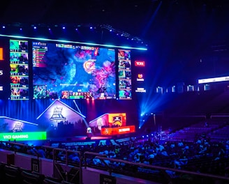 A large esports arena featuring a massive screen displaying a video game, surrounded by sponsor logos and team names. The auditorium is filled with spectators watching the gaming event. Neon blue and purple lights illuminate the stage, creating a vibrant atmosphere.