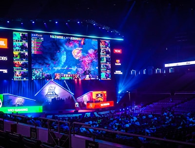 A large esports arena featuring a massive screen displaying a video game, surrounded by sponsor logos and team names. The auditorium is filled with spectators watching the gaming event. Neon blue and purple lights illuminate the stage, creating a vibrant atmosphere.