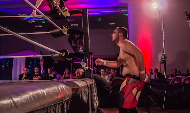 A wrestling match taking place in an indoor venue, where one participant is crouched on the top rope of the ring, while the other stands outside the ring looking at them. A crowd watches the event enthusiastically from their seats.