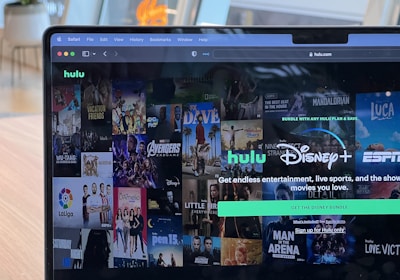 A laptop screen displays a web page featuring a mix of movie and TV show covers, highlighting popular titles and a Hulu and Disney+ bundle promotion. There are visible icons and images of films and shows alongside promotional text offering entertainment and live sports.