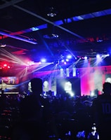 A dimly lit indoor arena filled with an audience. Bright stage lights in red and blue illuminate multiple large screens displaying graphics.