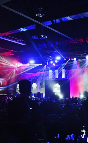 A dimly lit indoor arena filled with an audience. Bright stage lights in red and blue illuminate multiple large screens displaying graphics.