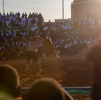 A vibrant wrestling match taking place outdoors, surrounded by a large crowd of spectators. The atmosphere is charged with excitement as people stand and sit around the dirt arena, intently watching the competitors. The sun is setting, casting a warm glow over the scene.