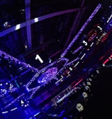 A large indoor arena with colorful and vibrant lighting, featuring a center stage surrounded by rigging and structures. Large numbered screens are visible, indicating marked sections from one to five. Various pieces of equipment and people are scattered around the venue.