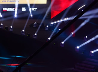 An array of bright stage lights beams across a dark arena, creating a dynamic atmosphere with a large screen displaying various logos and text. A camera crane is visible in the foreground, suggesting a professional event setup.