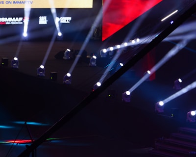 An array of bright stage lights beams across a dark arena, creating a dynamic atmosphere with a large screen displaying various logos and text. A camera crane is visible in the foreground, suggesting a professional event setup.
