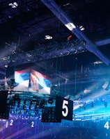 A large indoor arena is equipped with advanced lighting and audio systems. Suspended above the space are numerous digital displays showcasing numbers. The environment is filled with blue illumination and spotlights creating a vibrant atmosphere.