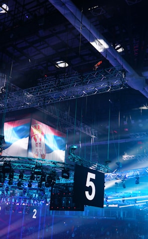 A large indoor arena is equipped with advanced lighting and audio systems. Suspended above the space are numerous digital displays showcasing numbers. The environment is filled with blue illumination and spotlights creating a vibrant atmosphere.