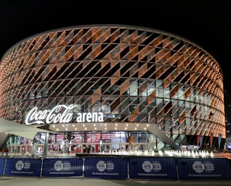A large, modern arena with a distinctive rounded design featuring a complex pattern of illuminated panels. The facade displays the 'Coca-Cola arena' signage prominently. The venue is surrounded by barriers with 'World Tennis League' branding. The lighting creates a vibrant and inviting atmosphere at night, with lighting around the entrance and inside the building.