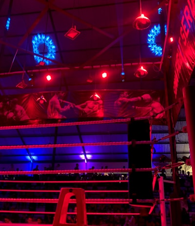 A brightly lit boxing or wrestling ring with vivid red and blue lights. Banners with images of fighters hang from the ceiling. The ropes of the ring are prominently visible, and a stool is placed inside the ring. The arena is filled with an audience, silhouetted against the colorful lighting.