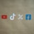 Four social media platform logos are arranged on a brown background. From left to right, the logos are for YouTube in red, TikTok in a combination of red, blue, and white, X in white, and Facebook in blue.