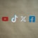 Four social media platform logos are arranged on a brown background. From left to right, the logos are for YouTube in red, TikTok in a combination of red, blue, and white, X in white, and Facebook in blue.