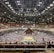 A large indoor arena packed with spectators seated in tiers around a central platform. The platform features traditional Japanese decor, indicating it may be used for sumo wrestling. The atmosphere is bustling, with people filling the seats and many spectators visible in the foreground.
