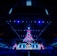 An expansive venue filled with a large audience, featuring a grand stage lit with vibrant blue and pink lights. The ceiling is decorated with a large display, and numerous screens showcase the event's branding. The atmosphere is dynamic and futuristic, reflecting a technologically advanced conference.